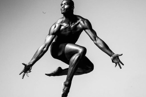 Black and white photo of Jamar Roberts jumping in the air with legs bent and arms stretched diagonally down to either side.
