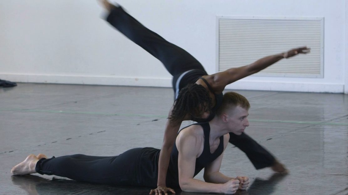 Image of two dancers. One is on floor, resting on forearms with stomach face down. Other dancer twists around and over him.