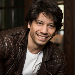 Herman Cornejo headshot. He is wearing a brown leather jacket and a white shirt. He has brown hair and brown eyes. He is leaning forward and smiling.