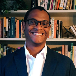 Paul J. Edwards headshot. He is wearing a black suit and white button down. He is standing in front of a bookcase. He is smiling, wearing glasses, and has short, dark hair.