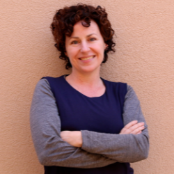 Mara Mills is standing in front of a plain, peach-colored wall. She has short, curly hair that is dark brown. She is smiling and standing with her arms crossed. She is wearing a blue long-sleeved shirt that is lighter on the sleeves and darker on the bodice. The photo is from the waist up.