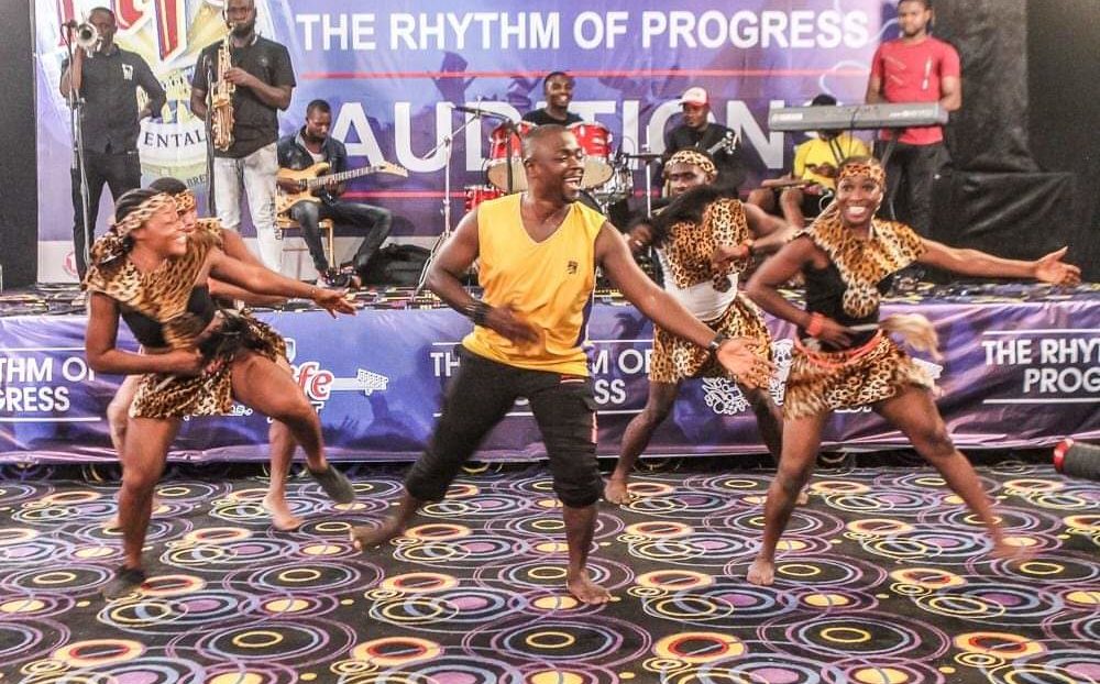 Nicholas Akas dances with a group. They are in a space with decorative walls and flooring. Akas is standing in the front. There are 4 people behind him. They are wearing a mix of attire with yellow and black colors.