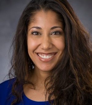 Photo of Sadye Paez wearing a blue shirt with a black background. She has brown wavy hair.