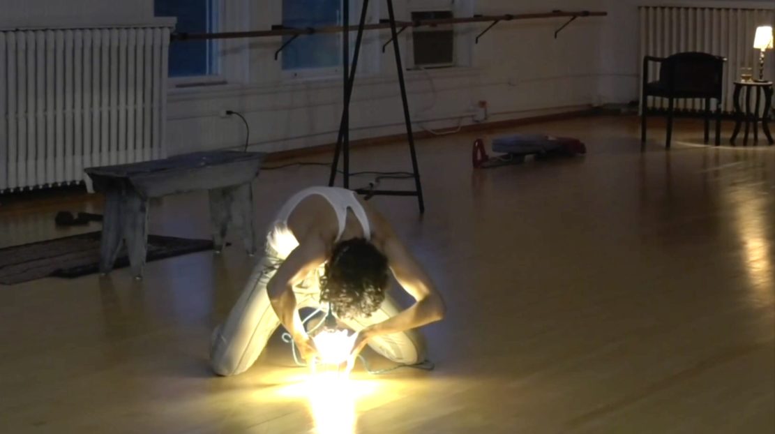 Kara Wilkes is dancing in a dance studio. She holds a ball of lights in a crouched position. She is wearing a white top and pants.