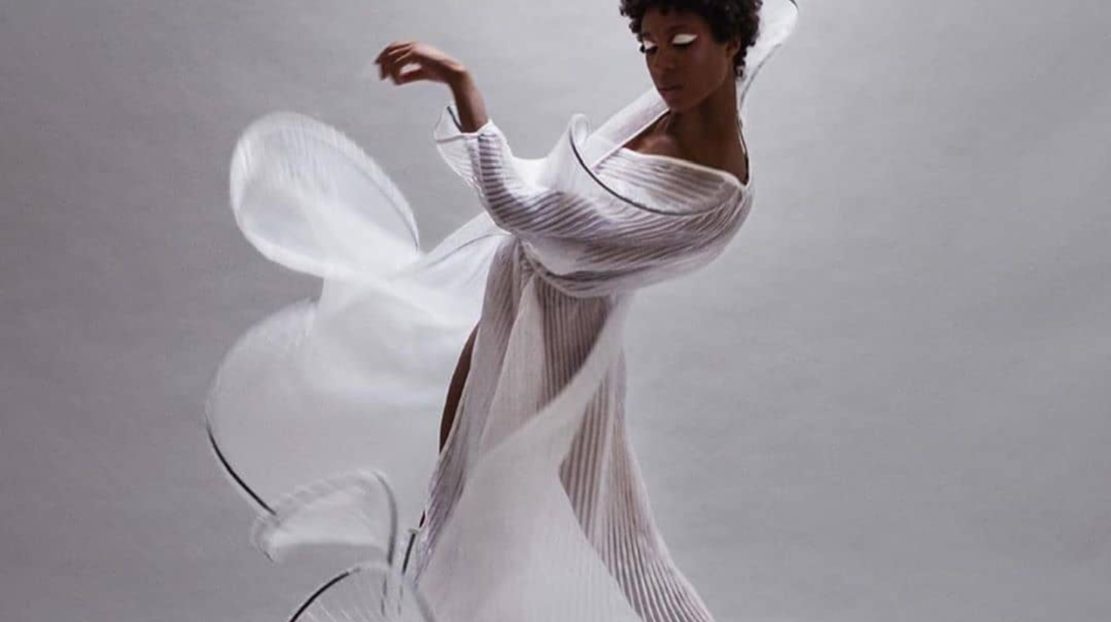 Adji Cissoko is in a dance pose with her feet and arms angled diagonally. One arm is above her head. She is wearing a sheer, white gown. She is in front of a gray backdrop.