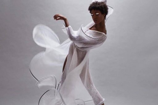 Adji Cissoko is in a dance pose with her feet and arms angled diagonally. One arm is above her head. She is wearing a sheer, white gown. She is in front of a gray backdrop.