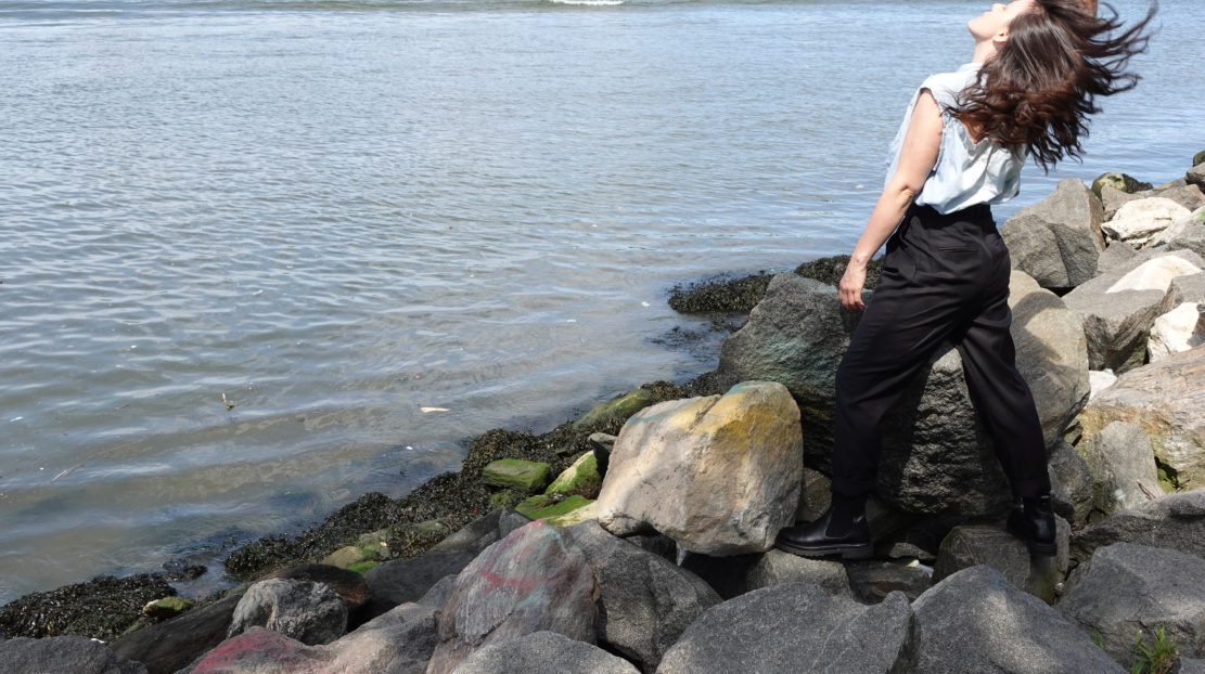 Emily stands in front of a body of water. She in on the right, wearing black pants and a light top. She has one arm above her head. She is standing on a rocky shore.