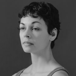 Headshot of Kara Wilkes in black and white. Kara stands in front of a black backdrop. She looks to the left with a soft expression. She has short dark hair and is wearing a leotard.