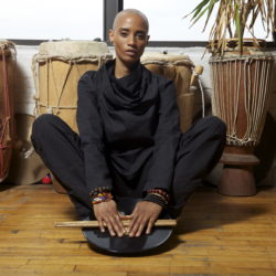 Val Jeanty sits in a cross-legged post in front of a set of drums. Val is holding a set of drum sticks.She has light hair that is very short, almost shaven. She is wearing all black.