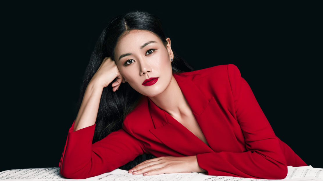 Jihye Lee is leaning on a table. She is wearing a red suit jacket, and has slicked-back black hair and red lips. She has a soft expression and is looking at the camera. The background is black.