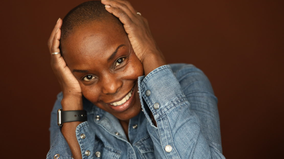 Hope Boykin, a Black woman with short dark hair, has her hands clasped on the sides of her head as she looks to the camera and smiles. She is wearing a chambray shirt, and apple watch, and a silver ring on her pinky finger.