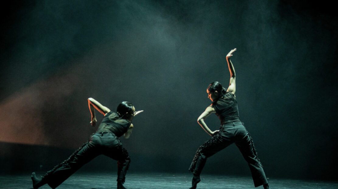 Ausia Jones, dancing with another dancer wearing black against a black background, surrounded by smoke.