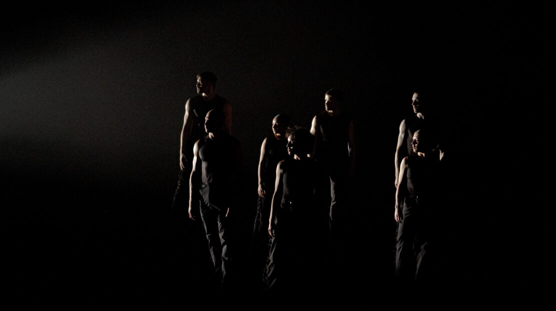 Ausia Jones, standing with other dancers, wearing all black against a black background. A single light shines from the left side.