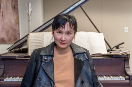 Head shot of Wang Lu, who has black hair with bangs in a ponytail and golden fair skin, sitting in front of piano.